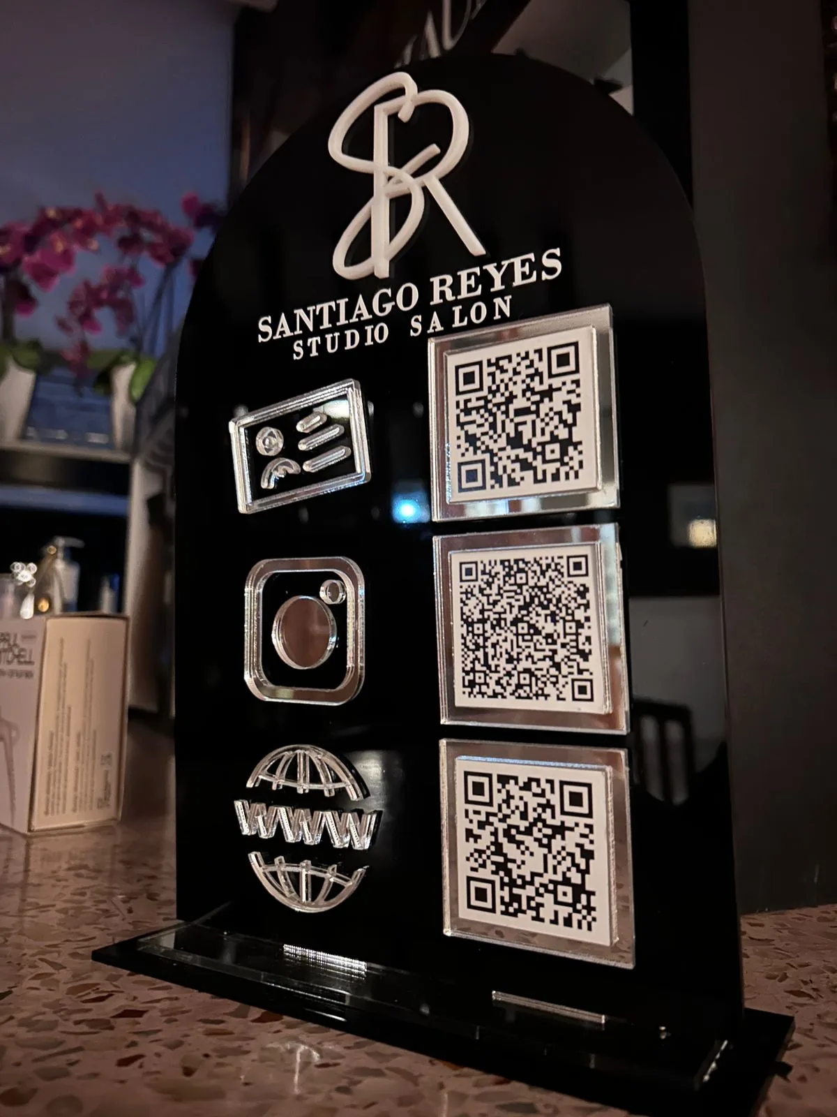 Display de mostrador en acrílico negro para Santiago Reyes Studio Salon, con monograma en relieve blanco y tres códigos QR en marcos de acrílico espejo plateado.