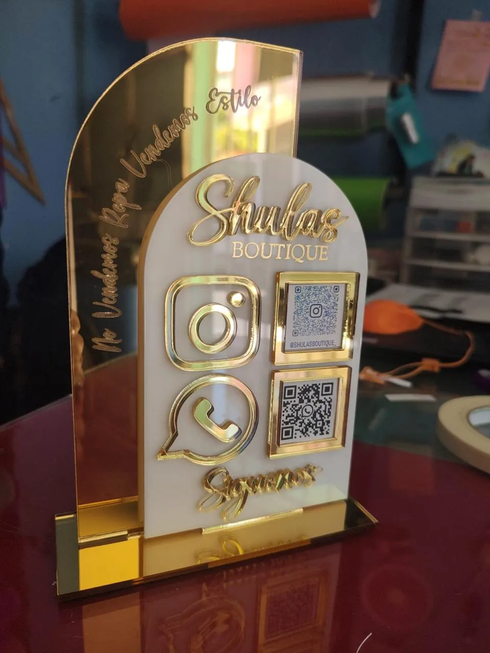 Display de mostrador en acrílico espejo dorado para Shulas Boutique, con logotipo en relieve, íconos de Instagram y WhatsApp y códigos QR grabados.