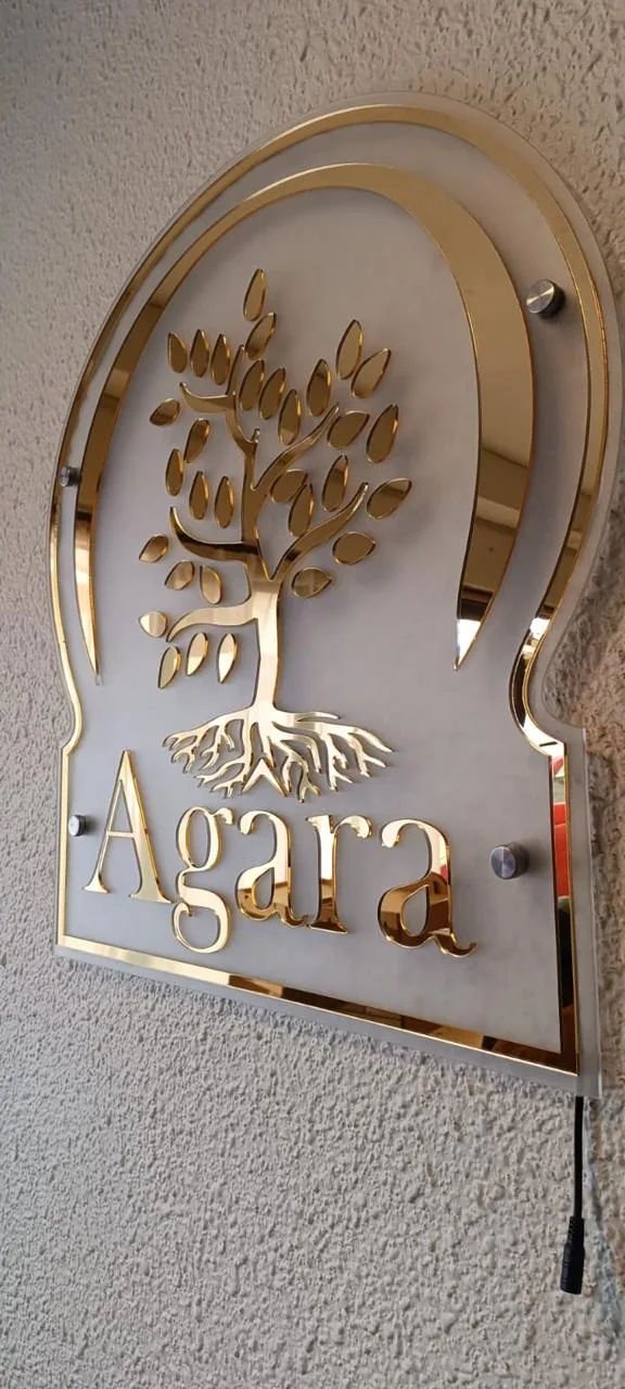 Letrero comercial de pared para la marca Agara en acrílico espejo dorado, con árbol de raíces grabado y montaje con separadores metálicos.