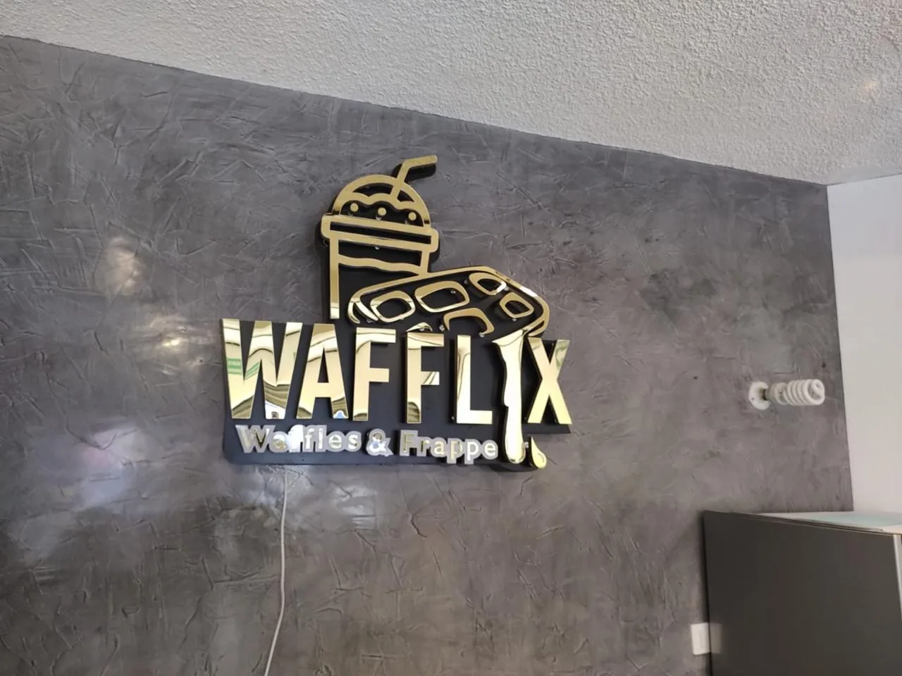 Letrero de pared para la wafflería Wafflix en acrílico espejo dorado y negro, con ilustración de frappé y waffles montada sobre muro de cemento pulido.