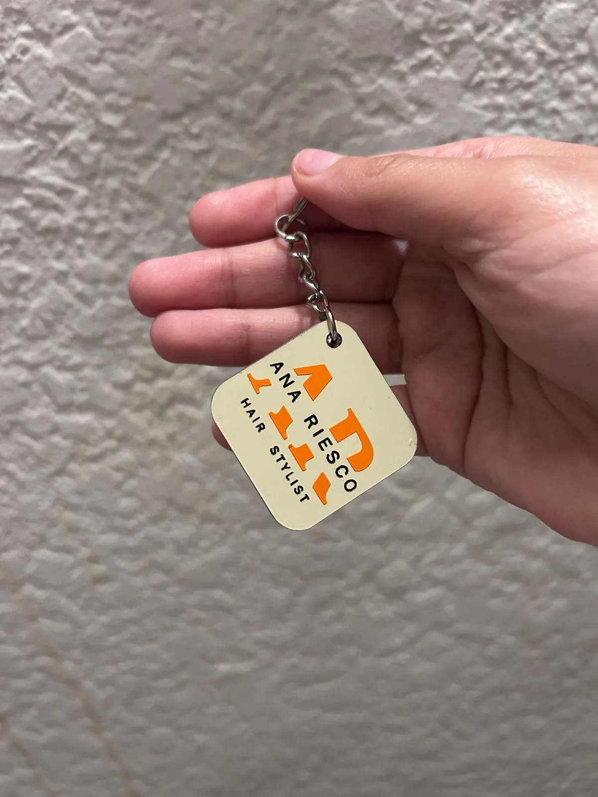 Llavero cuadrado personalizado con el logotipo naranja de Ana Riesco Hair Stylist, sostenido en la mano para mostrar su tamaño real.
